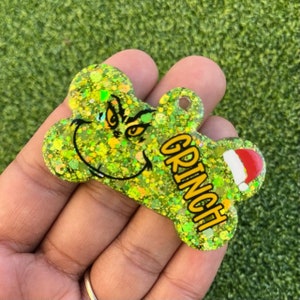 May include: Green glitter dog tag with a Grinch face and the word "GRINCH" in yellow letters. The tag has a red Santa hat on the side.