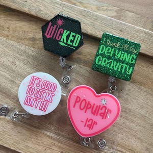 May include: Four different badge reel designs with glitter and text. The first is a black hexagonal badge reel with green glitter and the text "WICKED". The second is a green square badge reel with the text "I think I'll try defying gravity". The third is a white round badge reel with the text "It's good to see me isn't it!". The fourth is a pink heart-shaped badge reel with the text "Popular -lar".