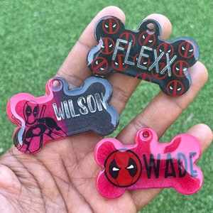 May include: Three bone-shaped dog tags with red and black Deadpool character designs. The tags are personalized with the names "Flexx", "Wilson", and "Wade".