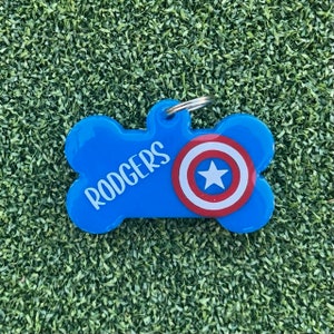 May include: A blue bone-shaped pet ID tag featuring the name RODGERS in white capital letters and a circular red, white, and blue shield graphic with a central white star. The tag includes a silver metal split ring attached to the top loop.