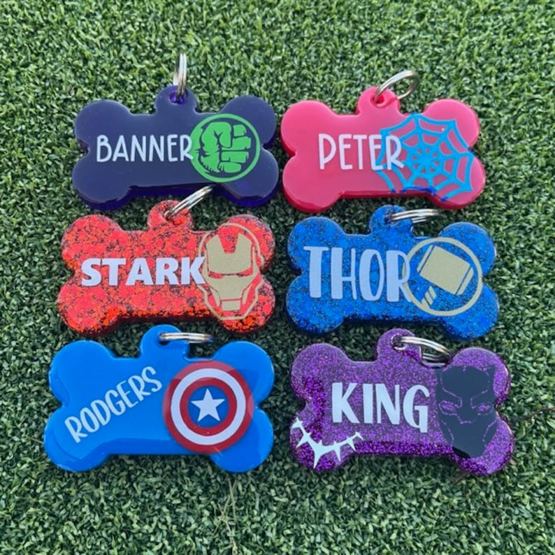 May include: Six bone-shaped pet ID tags featuring superhero themes. The tags are labeled Banner, Peter, Stark, Thor, Rodgers, and King, each with a corresponding graphic icon including a fist, spider web, mask, hammer, shield, and panther mask.