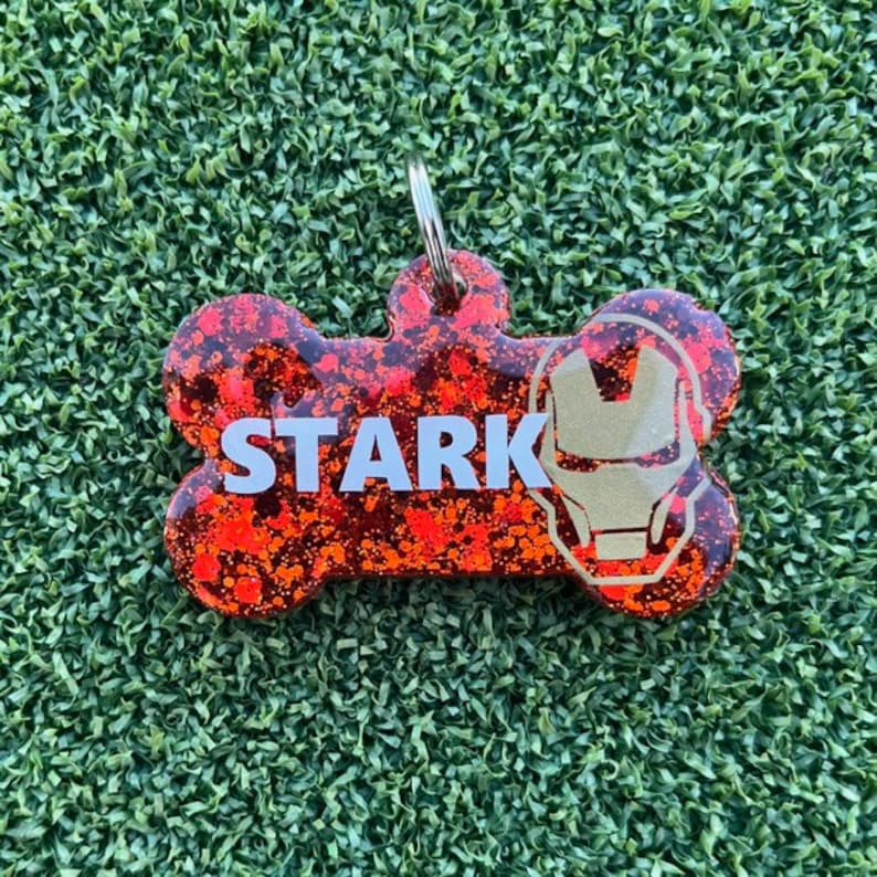May include: A bone-shaped pet ID tag featuring a red and black glittery finish. The word STARK is printed in white capital letters on the left side, and a gold-colored outline of a superhero helmet is positioned on the right side. A metal ring is attached.