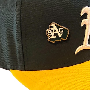 May include: A baseball cap with a dark green crown and a yellow brim. A gold-trimmed pin featuring the letters "A" and "S" in white and green is affixed to the cap. The letter "1" is embroidered in white.
