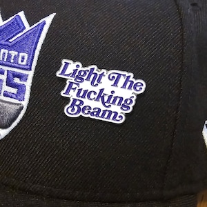 May include: A black baseball cap with a purple and white logo and a silver pin that says "Light The Fucking Beam" in purple letters.