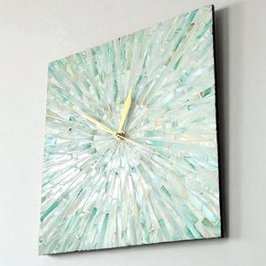 May include: Square wall clock with a mosaic design in shades of blue and white. The clock face has a sunburst pattern, with gold-coloured hands indicating the time. The clock is mounted on a white wall.