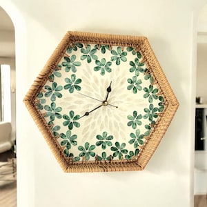 Könnte beinhalten: Eine sechseckige Wanduhr mit einem geflochtenen Rattanrahmen und einem weißen Zifferblatt mit grünem Blumendesign. Die Uhrzeiger sind schwarz und goldfarben.