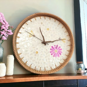 May include: A round, woven clock with a white face and a pink flower design. The clock face is made of small, white tiles.