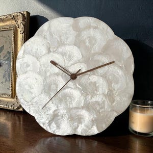 May include: A round wall clock featuring a face composed of overlapping white iridescent shell discs. The clock has three thin, dark brown metal hands centered on the face. The surface has a textured, pearlescent appearance with a glossy finish.