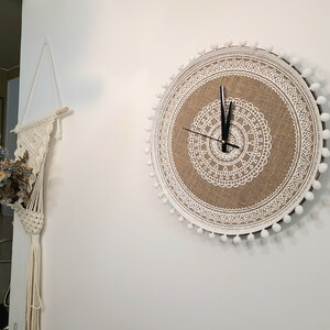 Peut inclure: Une horloge ronde en toile de jute avec un motif mandala blanc et une bordure de pompons blancs. Les aiguilles de l'horloge sont noires.