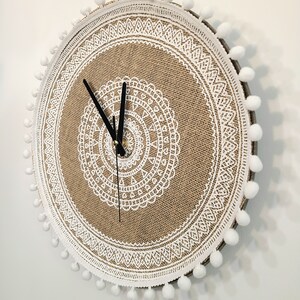 Peut inclure: Une horloge ronde en toile de jute avec un motif de dentelle blanche et des pompons blancs autour du bord.