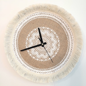 May include: A round burlap clock with a white lace design and a white fringe border. The clock has black hands and a white face.