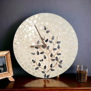 May include: A white round clock with a black and brown tree design. The clock has a white background with a pattern of small white circles. The tree design is made up of black leaves and brown branches.