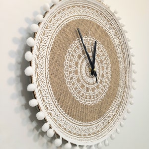 Peut inclure: Une horloge ronde en toile de jute avec un motif de dentelle blanche et des pompons blancs autour du bord. Les aiguilles de l'horloge sont noires.