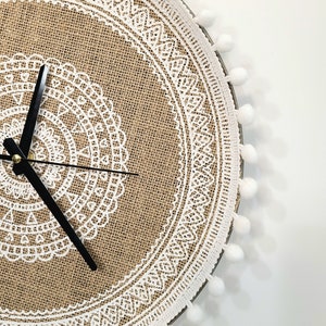 Peut inclure: Une horloge ronde en toile de jute avec un motif de dentelle blanche et un cadran noir. L'horloge est entour&eacute;e d'une bordure de pompons blancs.
