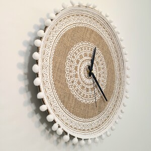 Peut inclure: Une horloge ronde en toile de jute avec une bordure en dentelle blanche et des pompons blancs autour du bord. Le cadran de l'horloge a un motif de mandala blanc.