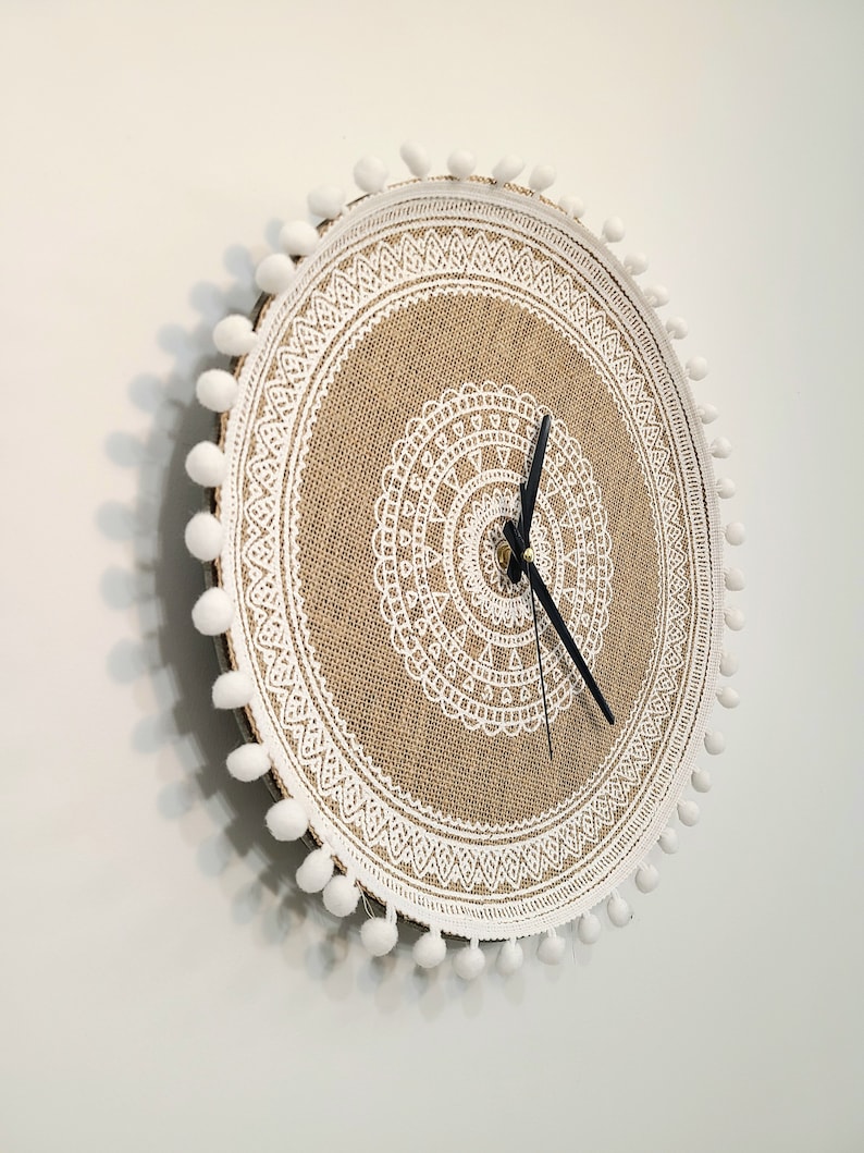 Peut inclure: Une horloge ronde en toile de jute avec une bordure en dentelle blanche et des pompons blancs autour du bord. Le cadran de l'horloge a un motif de mandala blanc.