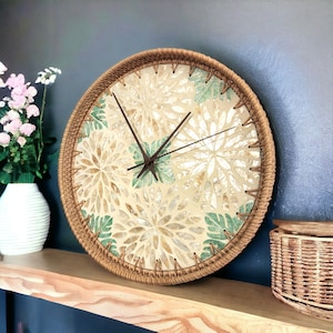 Puede incluir: Un reloj de pared redondo con un marco de ratán tejido y una esfera blanca con un diseño floral verde y dorado. Las manecillas del reloj son negras.