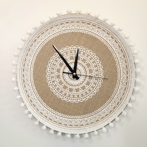 May include: A round burlap clock with a white lace design and white pom poms around the edge. The clock hands are black.