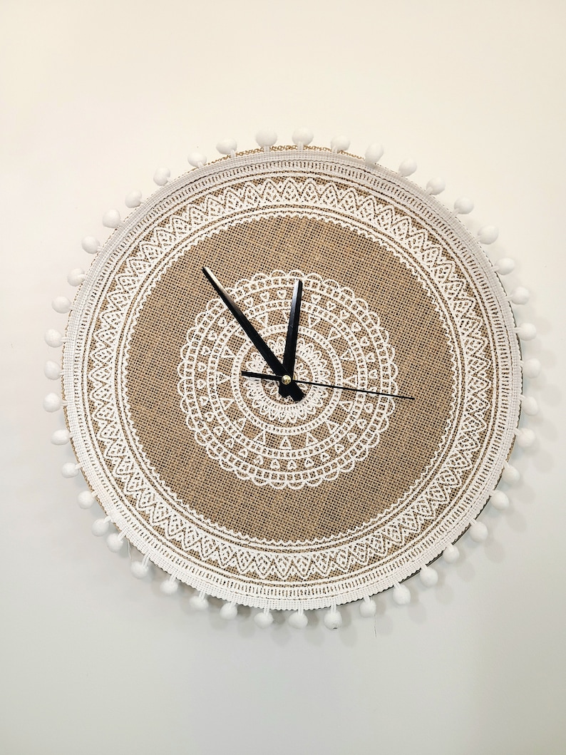Peut inclure: Une horloge ronde en toile de jute avec un motif de dentelle blanche et des pompons blancs autour du bord. Les aiguilles de l'horloge sont noires.