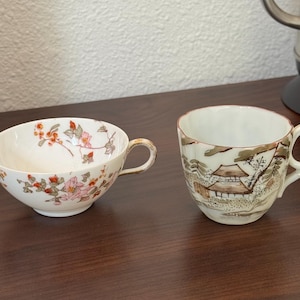 May include: Two porcelain teacups. One is white with pink and orange floral designs and a gold handle. The other cup has a landscape design with a brown and white color scheme.