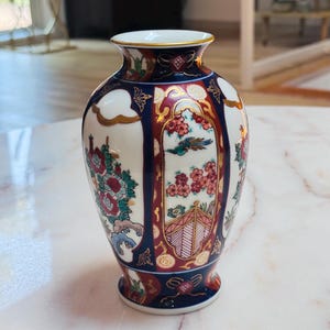 May include: A small porcelain vase featuring a white base with vertical panels in deep navy blue and dark red. The surface is decorated with intricate floral patterns, a bird, and architectural motifs, all accented with gold trim along the rim.