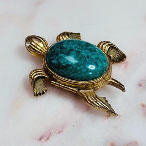 Könnte beinhalten: Eine goldfarbene Metallbrosche in Schildkrötenform mit einem großen, ovalen, gesprenkelten türkisfarbenen Cabochon-Stein als Panzer. Die Schildkröte hat strukturierte Gliedmaßen und einen kleinen, klaren, runden Strassstein am Kopf sowie ein Scharnier.