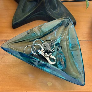 Puede incluir: Un plato triangular de vidrio color turquesa que contiene llaves y un dije decorativo. El plato tiene una cualidad ligeramente iridiscente. Las llaves están unidas a un llavero plateado.