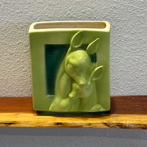 May include: A green ceramic vase with a rectangular shape. The vase features a relief of two deer, one embracing the other. The vase is a light green color with a darker green background.