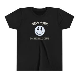 Può includere: T-shirt nera con la scritta "NEW YORK PICKLEBALL CLUB" in bianco. Una faccina sorridente azzurra con due fulmini è centrata sotto le parole "NEW YORK". La maglietta è realizzata in un materiale morbido.