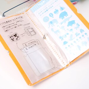 May include: A yellow binder with clear plastic pockets and a white sheet with blue and white illustrations of animals, a tent, a hot air balloon, a sun, and a flower. The text on the white sheet reads "パッケージごと、シートシール・スッキリ収納できます。"