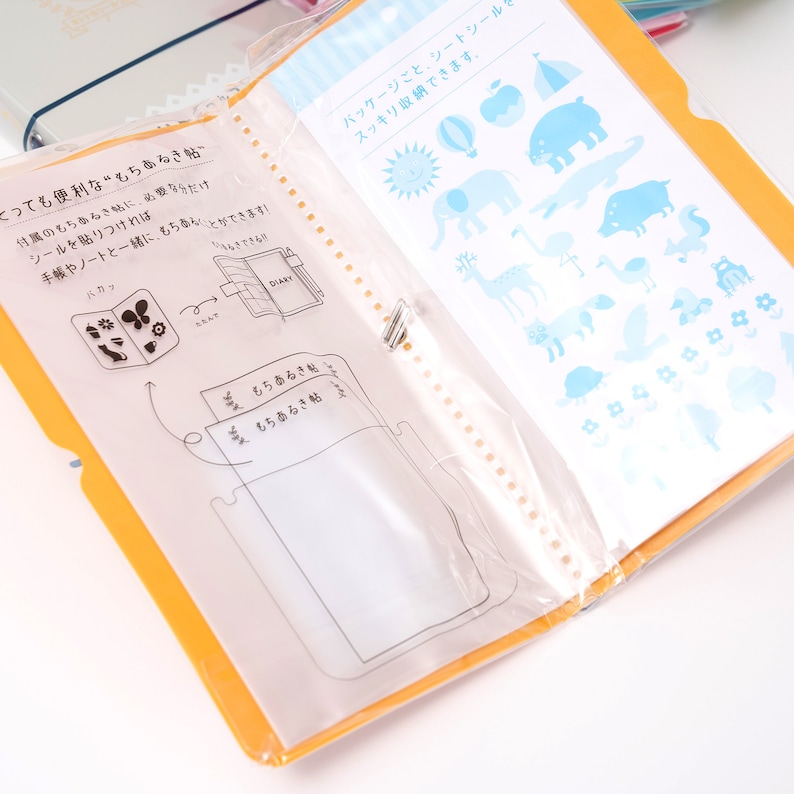 May include: A yellow binder with clear plastic pockets and a white sheet with blue and white illustrations of animals, a tent, a hot air balloon, a sun, and a flower. The text on the white sheet reads "パッケージごと、シートシール・スッキリ収納できます。"