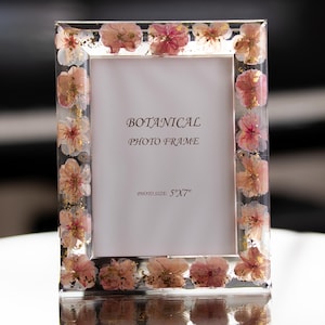 May include: Clear botanical photo frame with dried pink and white flowers embedded in the border. The frame is rectangular and features the text "BOTANICAL PHOTO FRAME" and "PHOTO SIZE: 5"X7"" in the center.