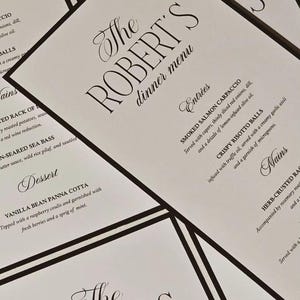 May include: A close-up of several printed menus. The menus are white with black text and a black border. The text reads "The ROBERT'S dinner menu" in elegant script. Menu items include Smoked Salmon Carpaccio and Crispy Risotto Balls.