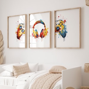 May include: Three watercolor prints featuring a colorful video game controller, headphones, and a second controller. The prints are framed in natural wood and hung on a white wall above a white daybed with a beige throw blanket.