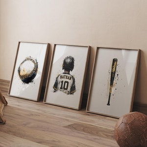 Puede incluir: Tres impresiones enmarcadas con temática de béisbol. La primera impresión presenta una pelota de béisbol con un diseño en blanco y negro. La segunda impresión presenta a un jugador de béisbol con una camiseta negra y dorada con el nombre "Nathan" y el número "10" en la espalda. La tercera impresión presenta un bate de béisbol con un diseño en negro y dorado.