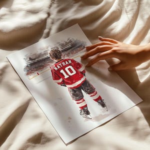 May include: A watercolor painting of a hockey player wearing a red jersey with the name "Nathan" and the number "10" on the back. The player is standing on the ice in front of a blurred background of a hockey arena.