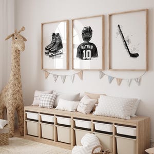May include: Three framed black and white illustrations of hockey equipment. The first illustration shows a pair of hockey skates, the second shows a hockey player wearing a jersey with the name "Nathan" and the number "10" on the back, and the third shows a hockey stick. The illustrations are hung on a white wall above a wooden bench with storage bins.