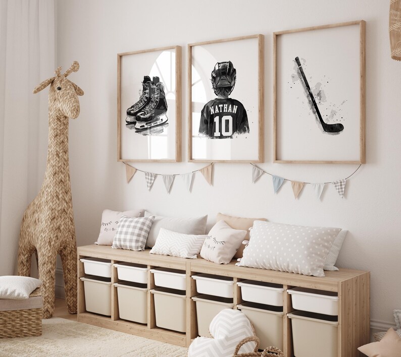 May include: Three framed black and white illustrations of hockey equipment. The first illustration shows a pair of hockey skates, the second shows a hockey player wearing a jersey with the name "Nathan" and the number "10" on the back, and the third shows a hockey stick. The illustrations are hung on a white wall above a wooden bench with storage bins.