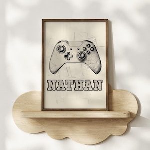 May include: A framed print featuring a sketch of a video game controller. The print includes the name "NATHAN" in bold, outlined letters. The artwork is displayed on a cloud-shaped wooden shelf.