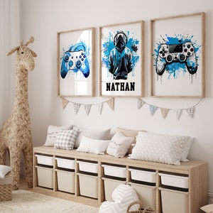 May include: Three framed prints with blue watercolor splashes and a gaming theme. The center print features a hooded figure with the name "NATHAN" below. The left and right prints feature a blue video game controller.