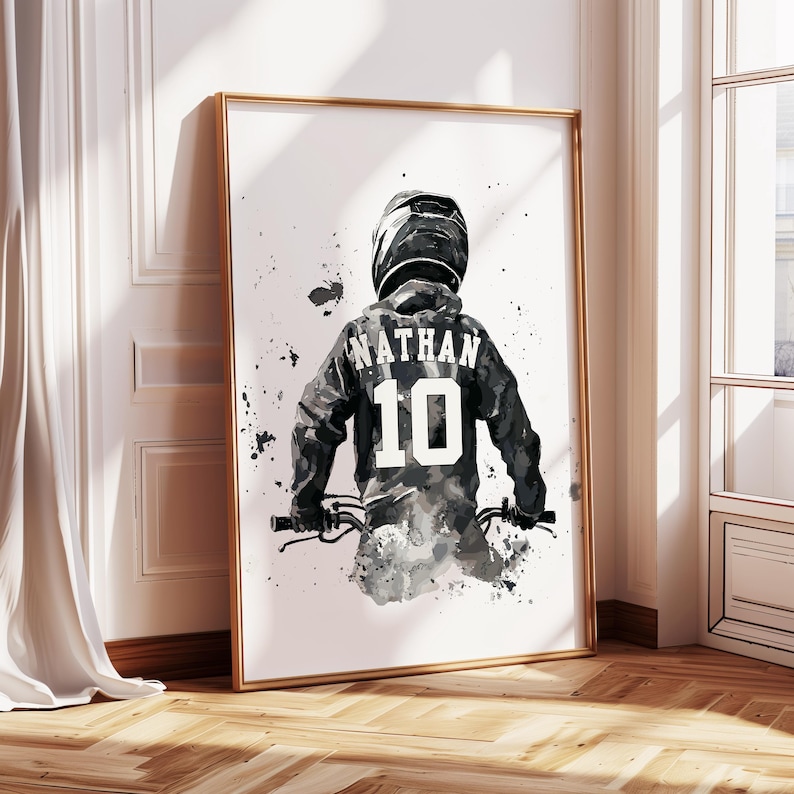 Peut inclure: Impression aquarelle en noir et blanc d'une personne portant un casque et une veste de motocross, avec le nom "NATHAN" et le num&eacute;ro "10" au dos. L'&oelig;uvre est encadr&eacute;e d'or et est pos&eacute;e contre un mur blanc.
