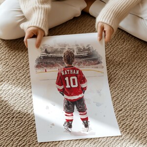 May include: A watercolor painting of a young hockey player wearing a red and white jersey with the name "Nathan" and the number "10" on the back. The player is standing on a hockey rink with a blurred background of a stadium.