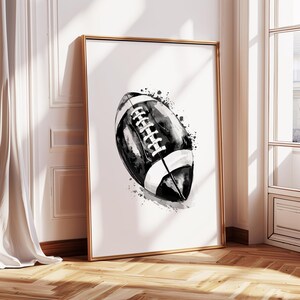 May include: Black and white watercolor illustration of an American football. The football is painted in a realistic style with details like laces and stitching. The background is a white canvas with subtle splashes of black paint.