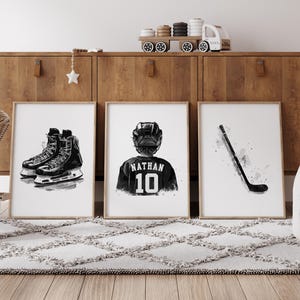 May include: Three black and white illustrations for a hockey themed nursery. The first image shows a pair of hockey skates, the second image shows a hockey player wearing a jersey with the name "Nathan" and the number "10" on the back, and the third image shows a hockey stick.