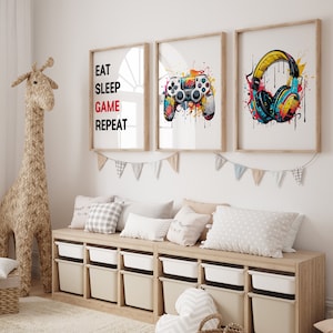 May include: Three framed prints with a gaming theme. The first print has the text "Eat Sleep Game Repeat" in black and red. The second print features a colorful illustration of a video game controller. The third print shows a pair of headphones with colorful paint splatters.