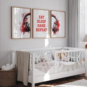 以下が含まれることがあります： ゲームをテーマにした3つのフレーム入りプリント。2つのプリントには、水彩画風のスプラッシュが付いた赤と黒のヘッドフォンが描かれています。中央のプリントには、赤い文字で「EAT SLEEP GAME REPEAT」と書かれています。プリントは白いベビーベッドのある部屋にあります。