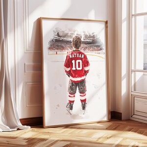 May include: A watercolor painting of a young hockey player wearing a red jersey with the name "Nathan" and the number "10" on the back. The player is standing on a white surface with a blurred image of a hockey arena in the background.