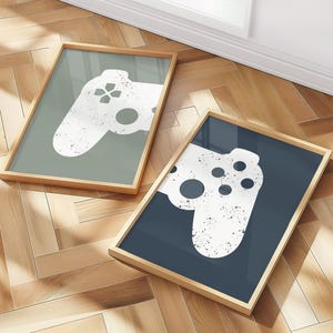 May include: Two framed prints featuring a white, distressed-style video game controller design. One print has a sage green background, the other a navy blue. Both are in light wood frames, resting on a herringbone wood floor.