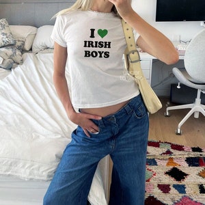 Może przedstawiać: Biała, krótka koszulka z napisem "I love Irish Boys" i zielonym sercem. Osoba nosi niebieskie dżinsy i białą torbę.