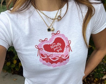 Puede incluir: Camiseta blanca recortada con un gráfico de pastel en forma de corazón rosa y rojo. El pastel tiene glaseado blanco y cerezas rojas. El texto en el pastel dice "I'm literally just a girl".